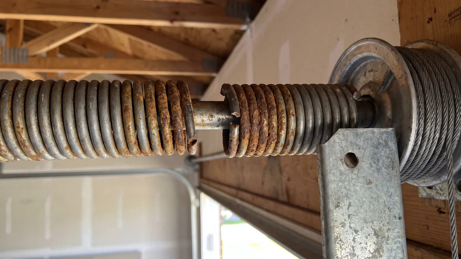 Broken torsion spring on the shaft above a residential garage door, the fracture rusted brown from salt air, with the cable drum beside it